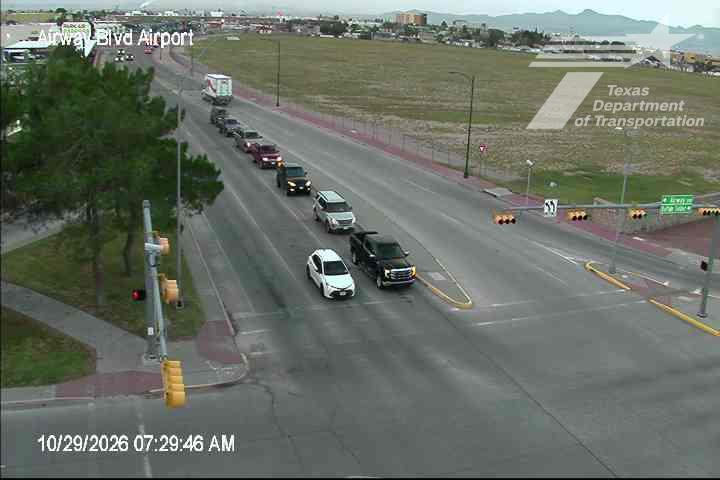 A line of vehicles, including a white sedan and a black pickup truck, waits at a traffic light at an intersection with a green highway sign indicating directions to the airport. The road is dry and clear with no visible incidents or construction.