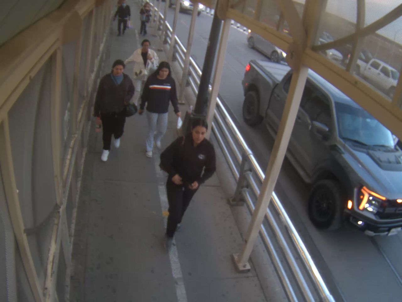 Pedestrians walk along a covered sidewalk adjacent to a road with light traffic, including a pickup truck and other vehicles.