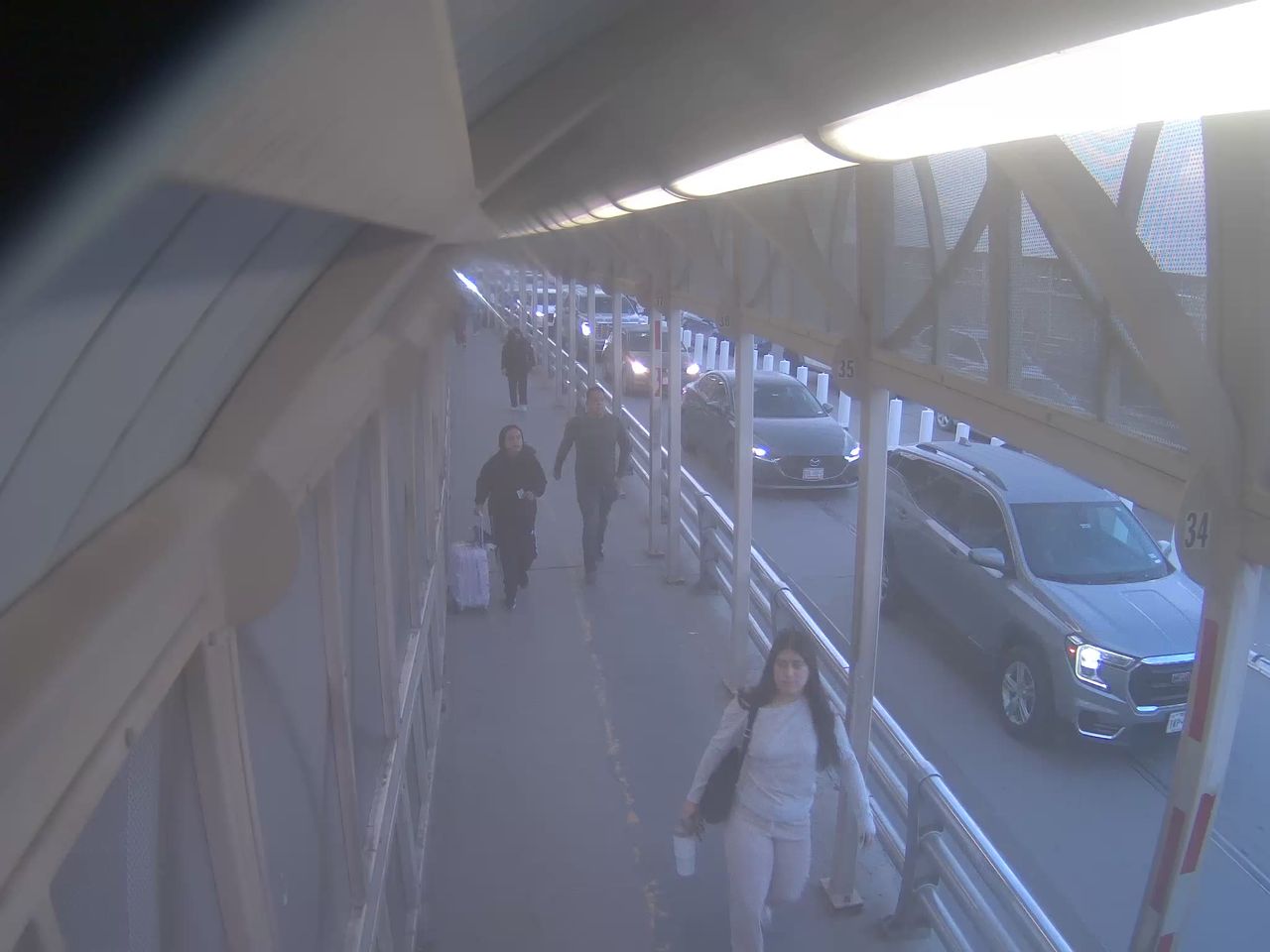 Pedestrians walk along a covered sidewalk adjacent to a roadway with light traffic; vehicles are moving slowly in both directions under an overpass structure.