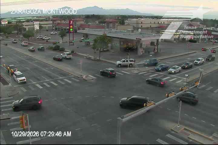 Vehicles navigate a multi-lane intersection with moderate traffic flow, passing a gas station and commercial buildings under clear skies.