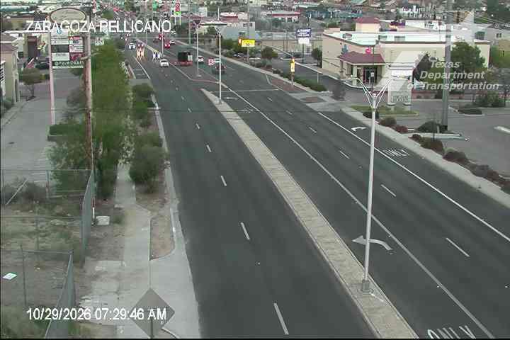 A wide, multi-lane road with light traffic moving in both directions, bordered by commercial buildings and sparse vegetation under clear skies.