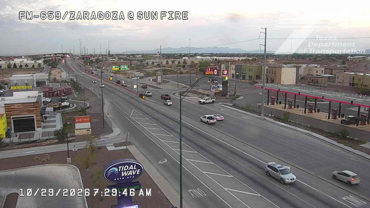 Light traffic flows along a multi-lane highway with commercial buildings and a gas station on the right, under a clear sky with distant mountains visible.