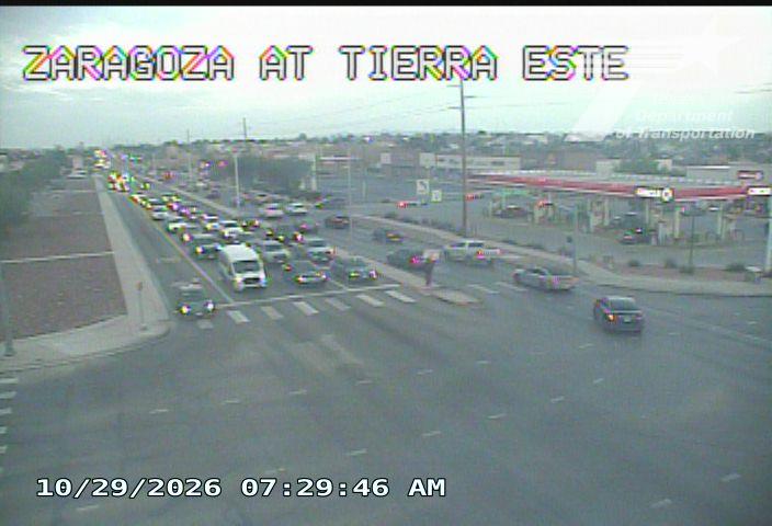 Traffic flows moderately at a multi-lane intersection with vehicles moving through crosswalks and passing a gas station; a lone pedestrian stands near the curb.