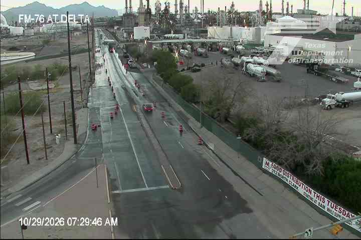 A wet multi-lane highway with light traffic flows past an industrial complex, with construction cones and barriers indicating a work zone; a red vehicle is visible in the center lane.