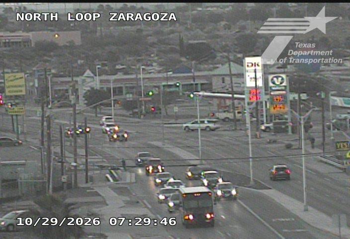 Traffic flows moderately at an urban intersection with a bus and multiple cars moving through the crossroads; a gas station is visible on the right side of the road.