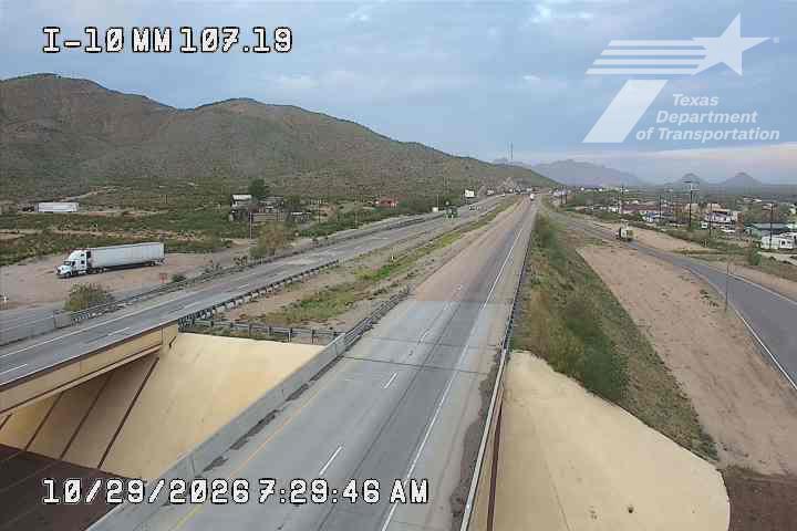A multi-lane highway with light traffic flows under an overpass, flanked by arid hills and sparse roadside development; a single semi-truck is parked on the shoulder, and no active road work or incidents are visible.