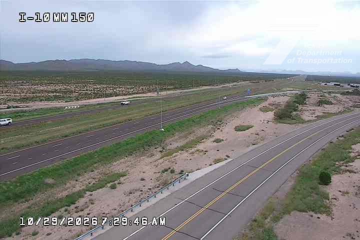 A wide highway stretches through a desert landscape with distant mountains under an overcast sky, showing light traffic with a few vehicles moving freely.