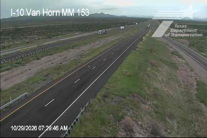 A wide, multi-lane highway stretches through a dry, arid landscape with sparse vegetation and distant mountains under an overcast sky; light traffic moves steadily in both directions with no visible incidents or road work.