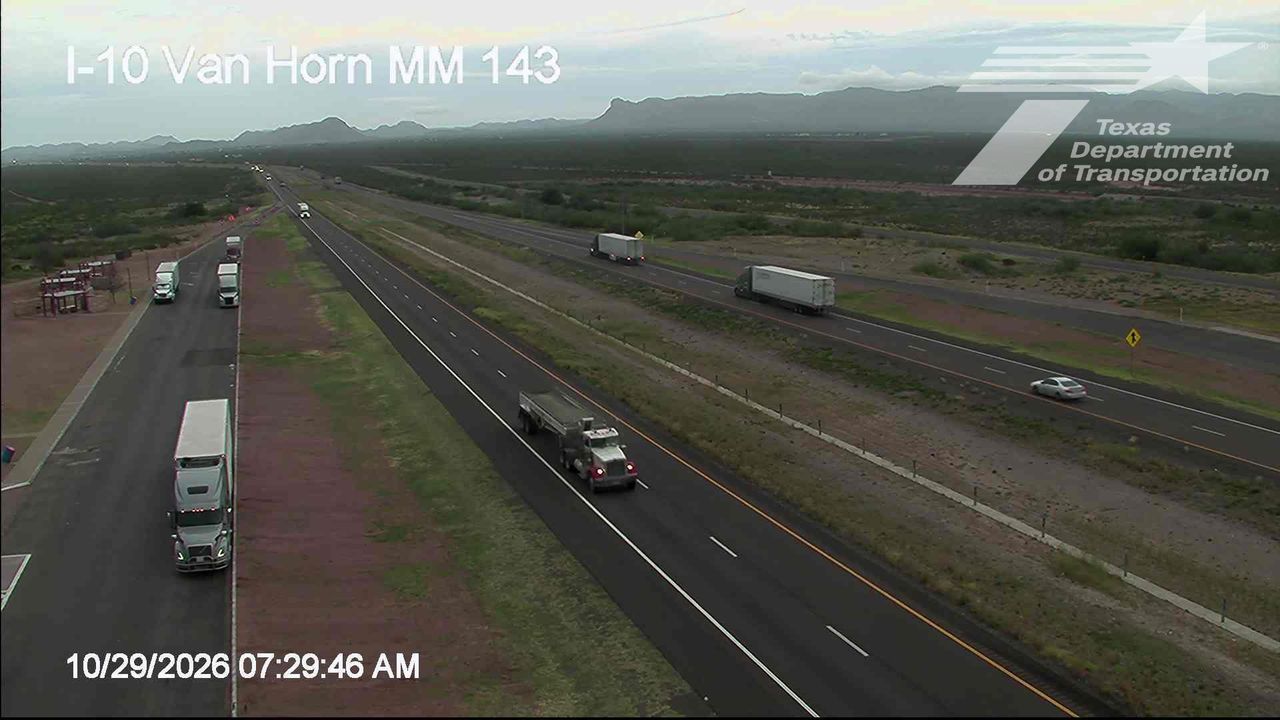 Multi-lane highway with moderate traffic flow, featuring several semi-trucks and a single white sedan; no visible incidents or road work zones; distant mountains under overcast skies.