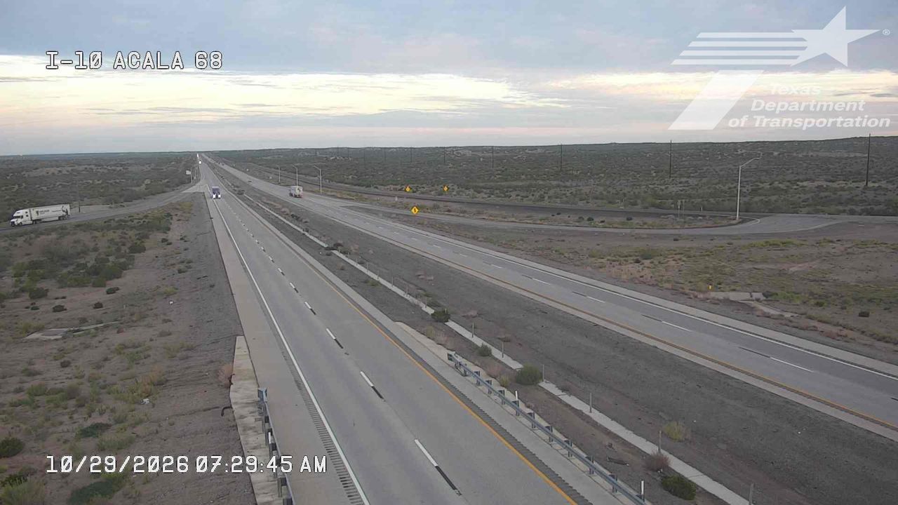 A wide, multi-lane highway stretches through a dry, arid landscape with sparse vegetation, showing light traffic with a few trucks moving steadily in both directions under a partly cloudy sky.