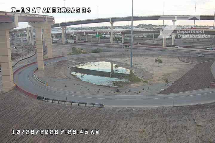 A multi-level highway interchange with a central traffic circle featuring standing water, surrounded by underdeveloped roadways and overpasses with minimal vehicle traffic.