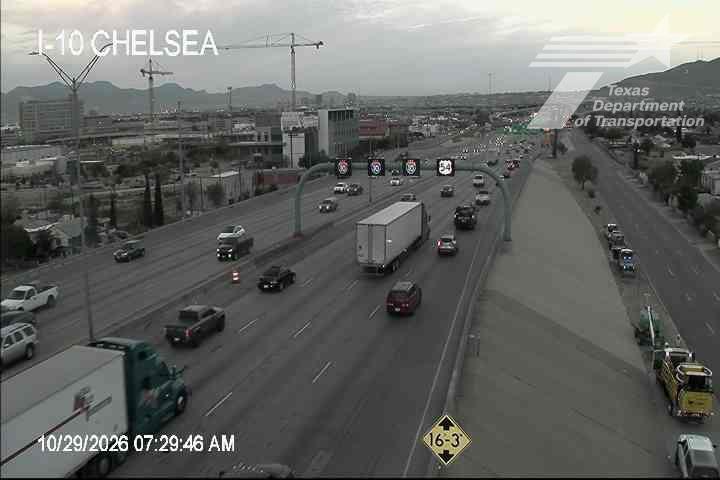 Multi-lane highway with moderate traffic flow, including semi-trucks and passenger vehicles, under overcast skies with distant mountains in view.