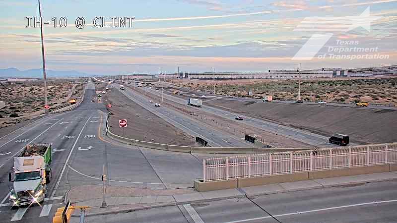 A wide highway intersection with light traffic, featuring a green dump truck at the curb and multiple vehicles moving through the lanes under clear skies.