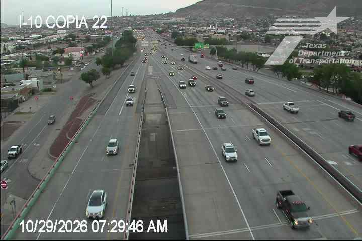 Multi-lane highway with moderate traffic flowing in both directions under overcast skies, featuring a central overpass structure and distant urban development.