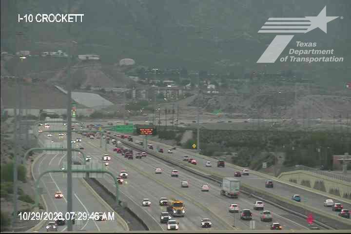 Moderate traffic flows along a multi-lane highway with a mix of cars and a semi-truck, under overcast skies with distant hills in the background.