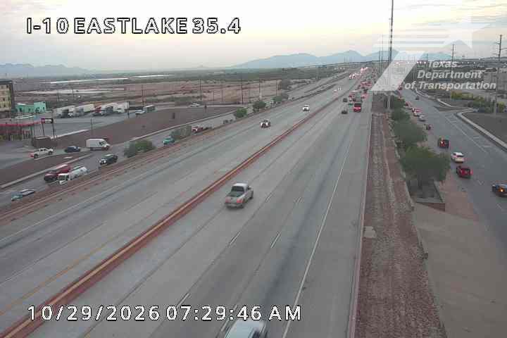 Light traffic flows along a multi-lane highway with vehicles moving at moderate speeds, including a pickup truck and several cars, under clear skies with distant mountains visible.