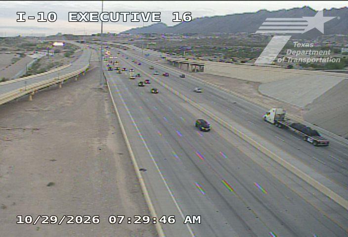 Multi-lane highway with moderate traffic flow, including a semi-truck and several cars, under clear skies with distant mountains in the background.
