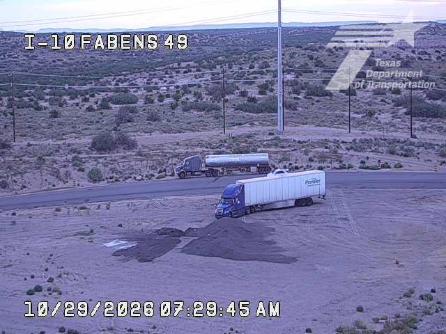 A semi-truck with a white trailer is parked on the shoulder of a desert highway, while another tanker truck is visible further down the road; no active traffic or work zones are present.