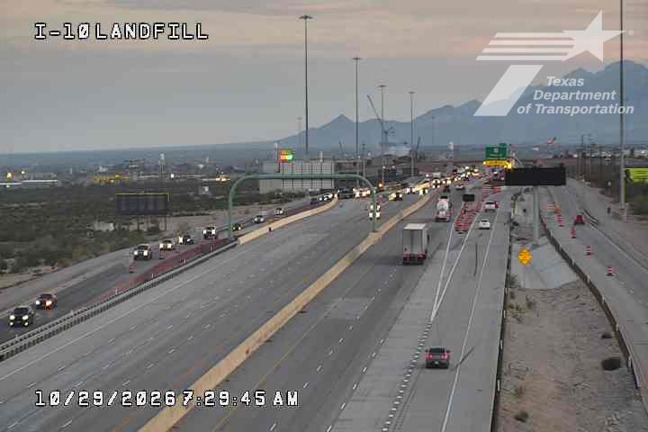 Multi-lane highway with moderate traffic flow, including trucks and cars, under clear skies with distant mountains in view; construction cones and barriers indicate active road work on the right side.