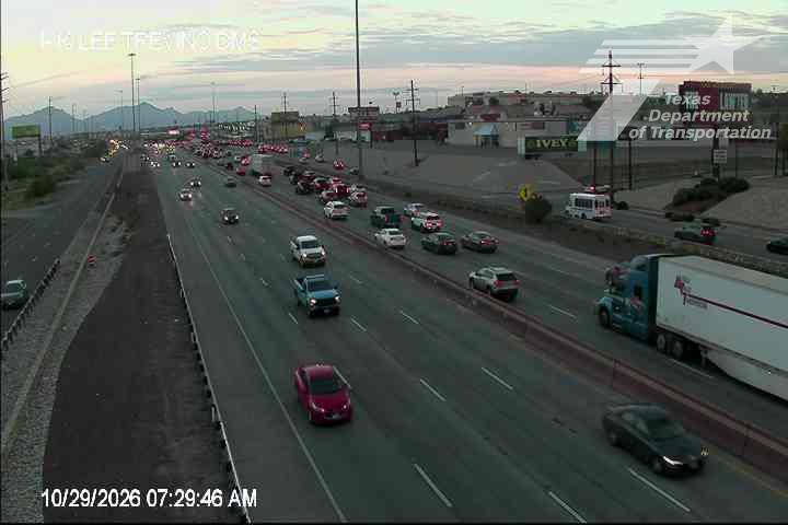 Multi-lane highway with moderate traffic flow during early morning hours, featuring a mix of passenger cars and a large semi-truck, with distant mountains under a soft sky.