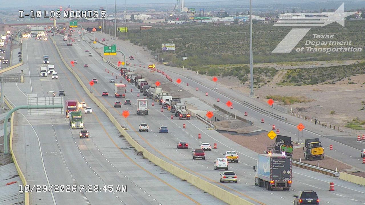Multi-lane highway with moderate traffic flow, including trucks and cars, near an active road construction zone marked by cones and barriers; workers are visible on foot near equipment.