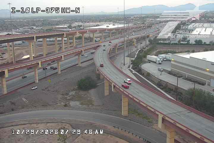 Multi-level highway interchange with moderate traffic flow under clear skies, featuring overpasses, commercial buildings, and distant mountains.