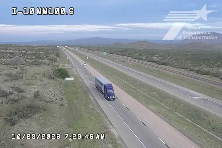 A blue semi-truck travels along a multi-lane highway with light traffic, surrounded by open grassland and distant hills under a partly cloudy sky.