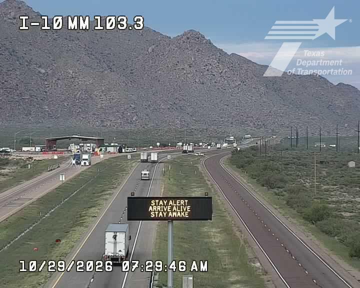 Free-flowing traffic on a multi-lane highway with trucks and cars moving under a clear sky, bordered by arid mountains and sparse vegetation.