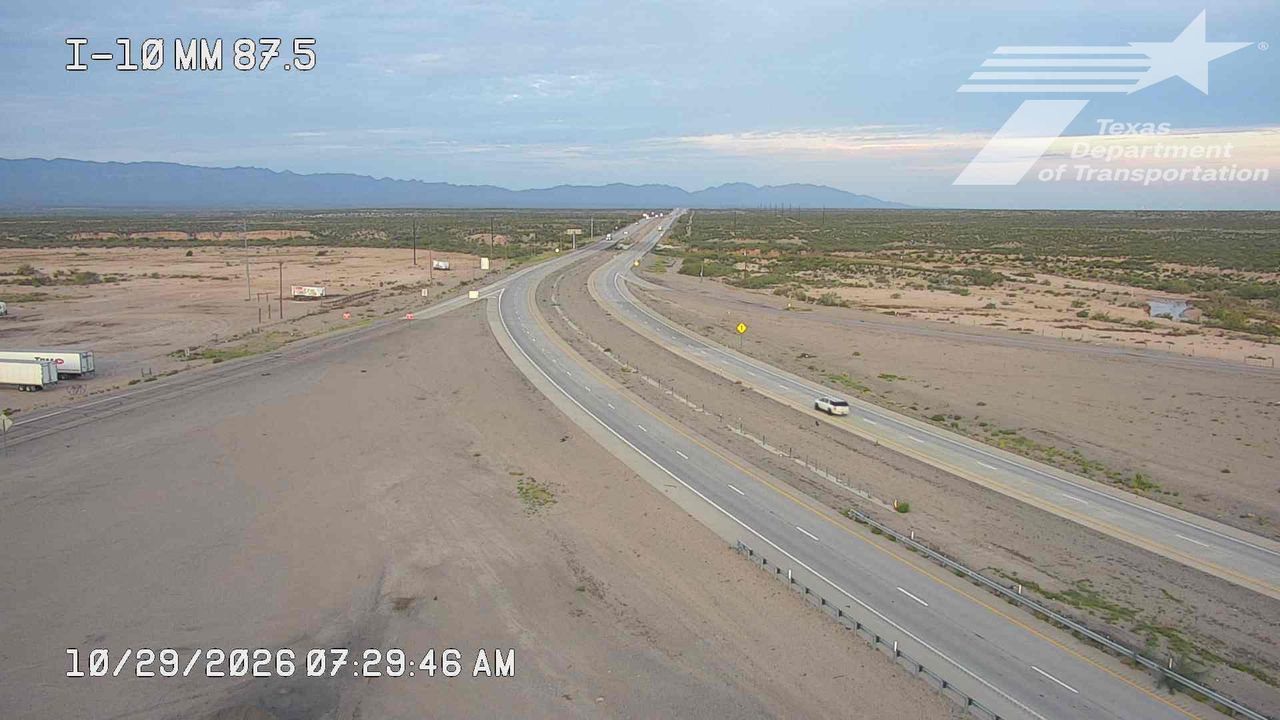 A wide, multi-lane highway curves through a dry, arid landscape with distant mountains under a clear sky; light traffic moves steadily along the road with no visible incidents or road work.