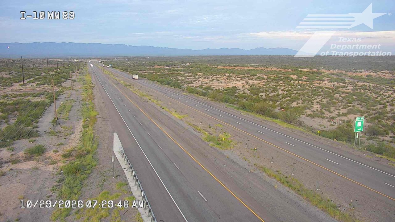 A wide, multi-lane highway stretches through a dry, arid landscape with sparse vegetation and distant mountains under a clear sky; light traffic moves steadily in both directions with no visible incidents or road work.