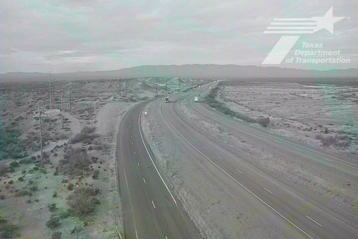 A multi-lane highway curves through a dry, arid landscape with sparse vegetation and distant hills under an overcast sky; light traffic moves steadily in both directions with no visible incidents or road work.