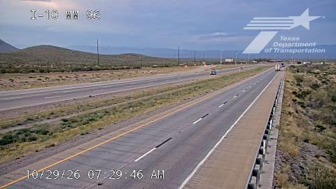 A multi-lane highway stretches through a dry, arid landscape with distant mountains under a partly cloudy sky; light traffic moves steadily in both directions with no visible work zones or incidents.