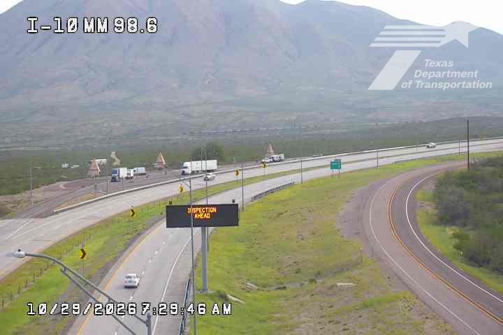 Multi-lane highway with moderate traffic flowing in both directions, featuring a work zone with cones and barriers, and a digital sign indicating an inspection ahead; distant mountains loom under clear skies.