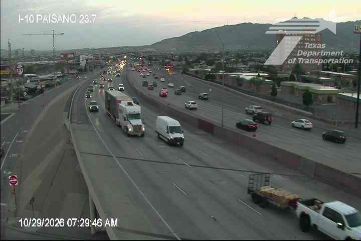 Multi-lane highway with moderate traffic flow, featuring semi-trucks, vans, and passenger cars moving in both directions under clear skies with distant hills in the background.
