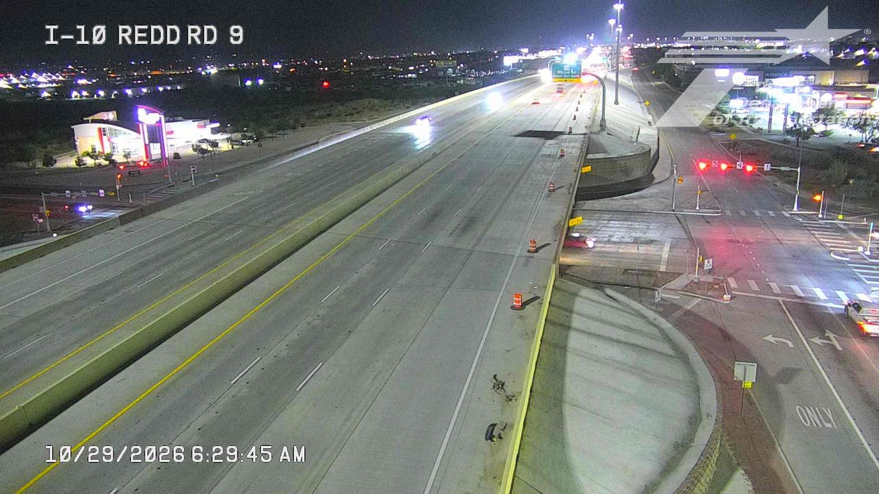 Nighttime view of a multi-lane highway with minimal traffic, illuminated by streetlights and commercial signage; a few vehicles are visible on the road and at an intersection with red signals, while construction cones line the shoulder near a work zone.