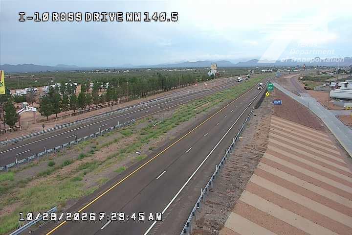Highway with light traffic moving in both directions, bordered by sparse vegetation and distant mountains under an overcast sky.