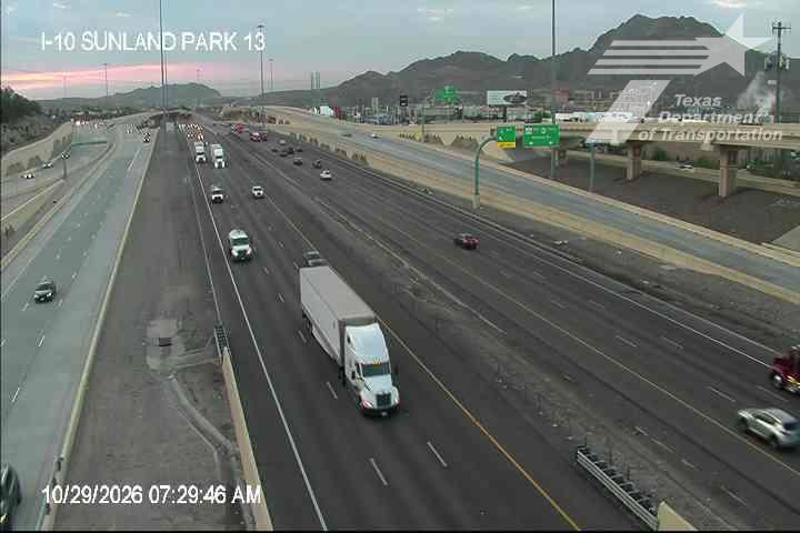 Multi-lane highway with moderate traffic flow, featuring a prominent semi-truck and several passenger vehicles moving in both directions under clear skies with distant mountains in view.