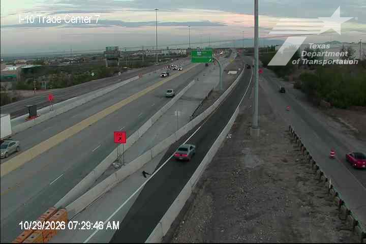 Multi-lane highway with moderate traffic flow, separated by concrete barriers; a lone vehicle is on the shoulder, and construction cones line the right lane; clear sky with soft lighting suggests early morning.