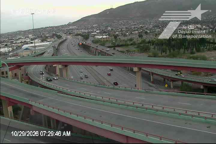 Multi-level highway interchange with moderate traffic flow under overcast skies, featuring green and red painted overpasses and distant urban development.