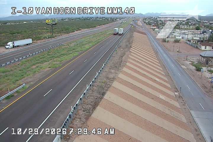Multi-lane highway with light traffic, featuring semi trucks and a wide median strip with striped pattern; distant mountains and clear skies indicate dry, sunny weather.