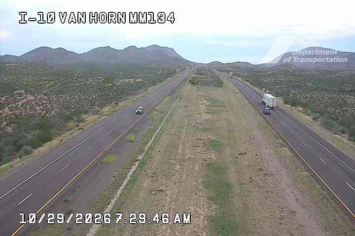 A wide highway stretches through a desert landscape with distant mountains, featuring light traffic with a few vehicles moving in both directions.