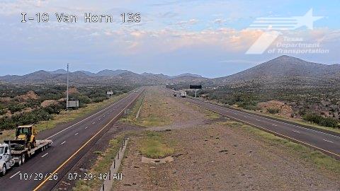 A wide highway stretches through a desert landscape with distant mountains, featuring light traffic and a large flatbed truck carrying heavy equipment on the left lane.