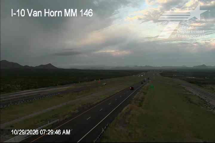 Light traffic on a multi-lane highway with distant mountains under a partly cloudy sky, with a few vehicles visible in both directions.