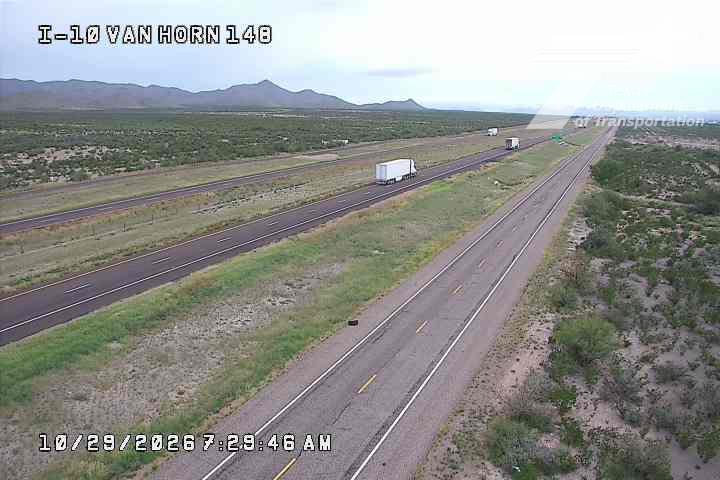 A multi-lane highway stretches through a desert landscape with distant mountains, featuring several semi-trucks traveling in both directions under clear skies.