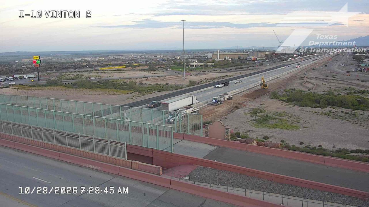 Highway under construction with light traffic and active road work, including trucks and heavy equipment on the right side of the road.