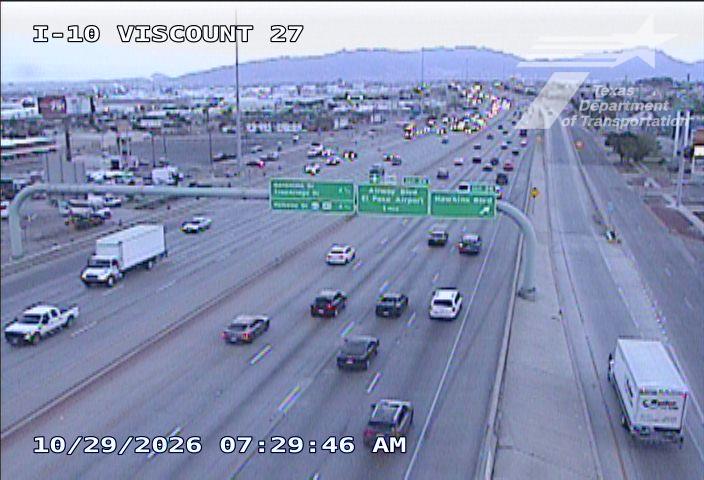 Multi-lane highway with moderate traffic flow, including passenger cars and trucks, under clear skies with distant hills in the background.