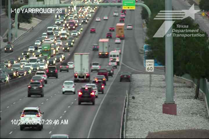 Heavy traffic flows in both directions on a multi-lane highway with numerous cars and trucks, including semi-trucks, under clear weather conditions.