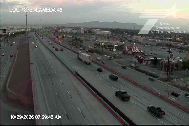 Multi-lane highway with moderate traffic flow, including trucks and passenger cars, under clear skies with distant mountains visible.