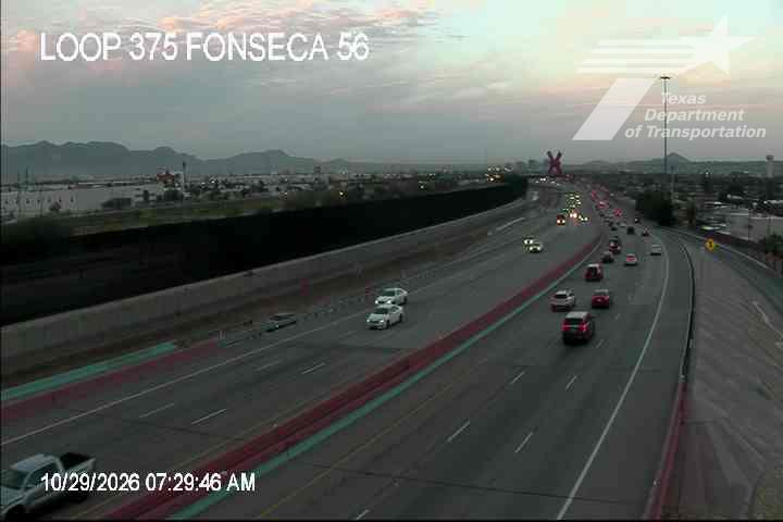 Light traffic flows along a multi-lane highway with distant mountains under a clear sky, featuring a mix of passenger cars and a pickup truck.