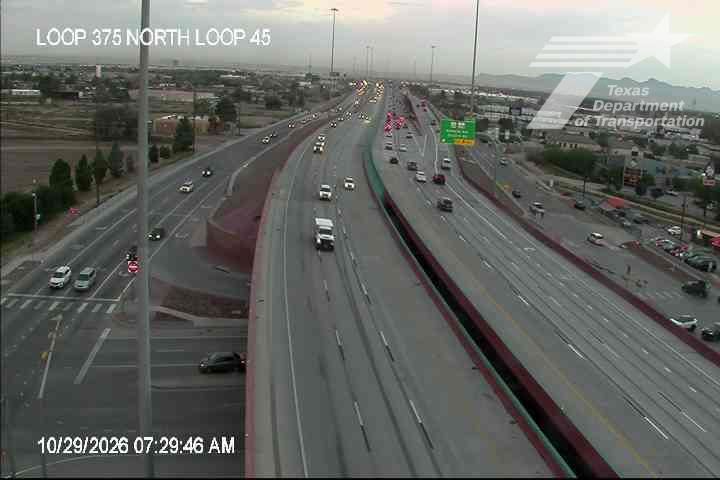 Multi-lane highway with moderate traffic flowing in both directions under clear skies, featuring an overpass and distant urban landscape.
